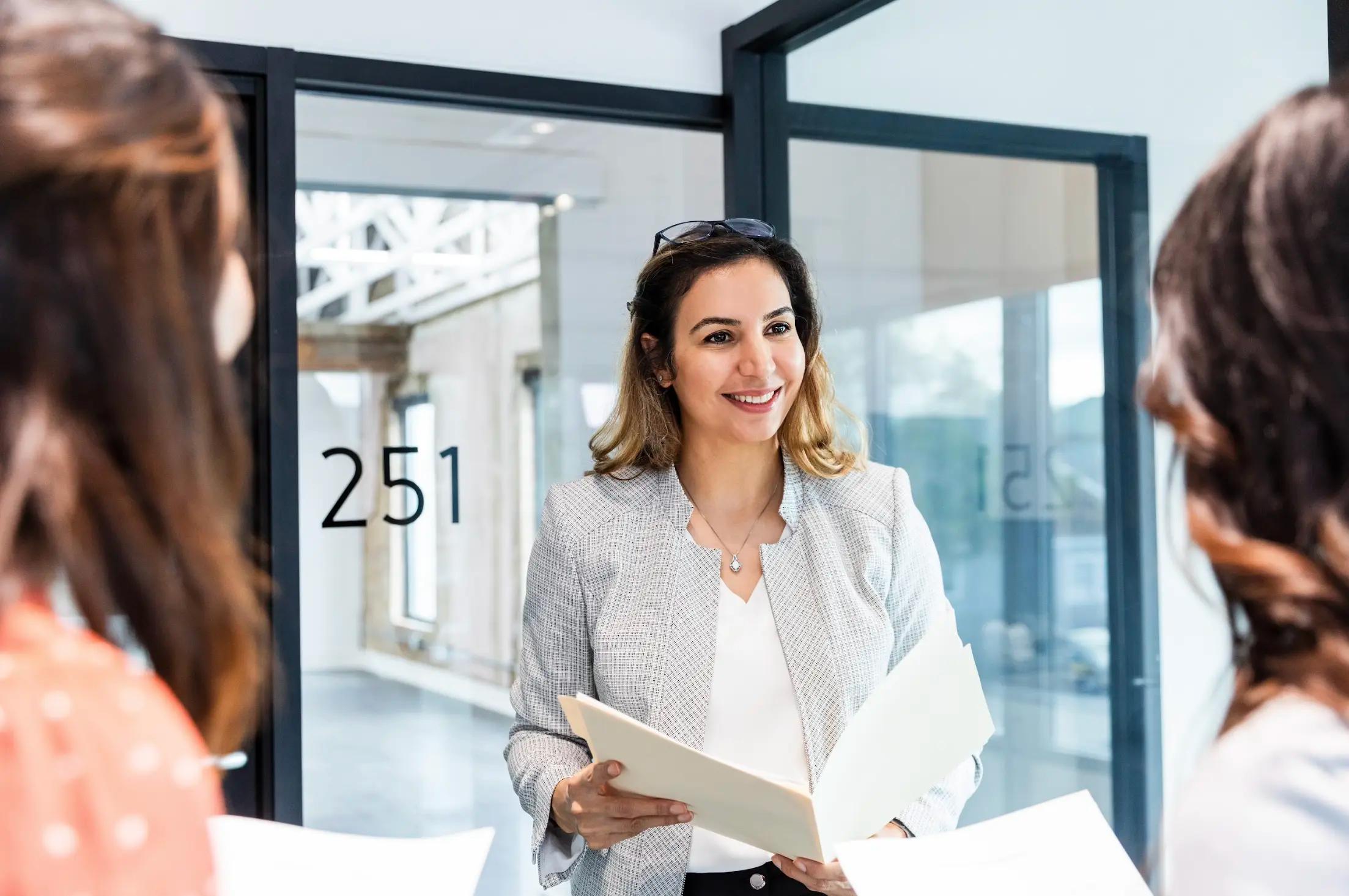Smiling woman holding a folder, speaking with two colleagues in a modern office hallway.