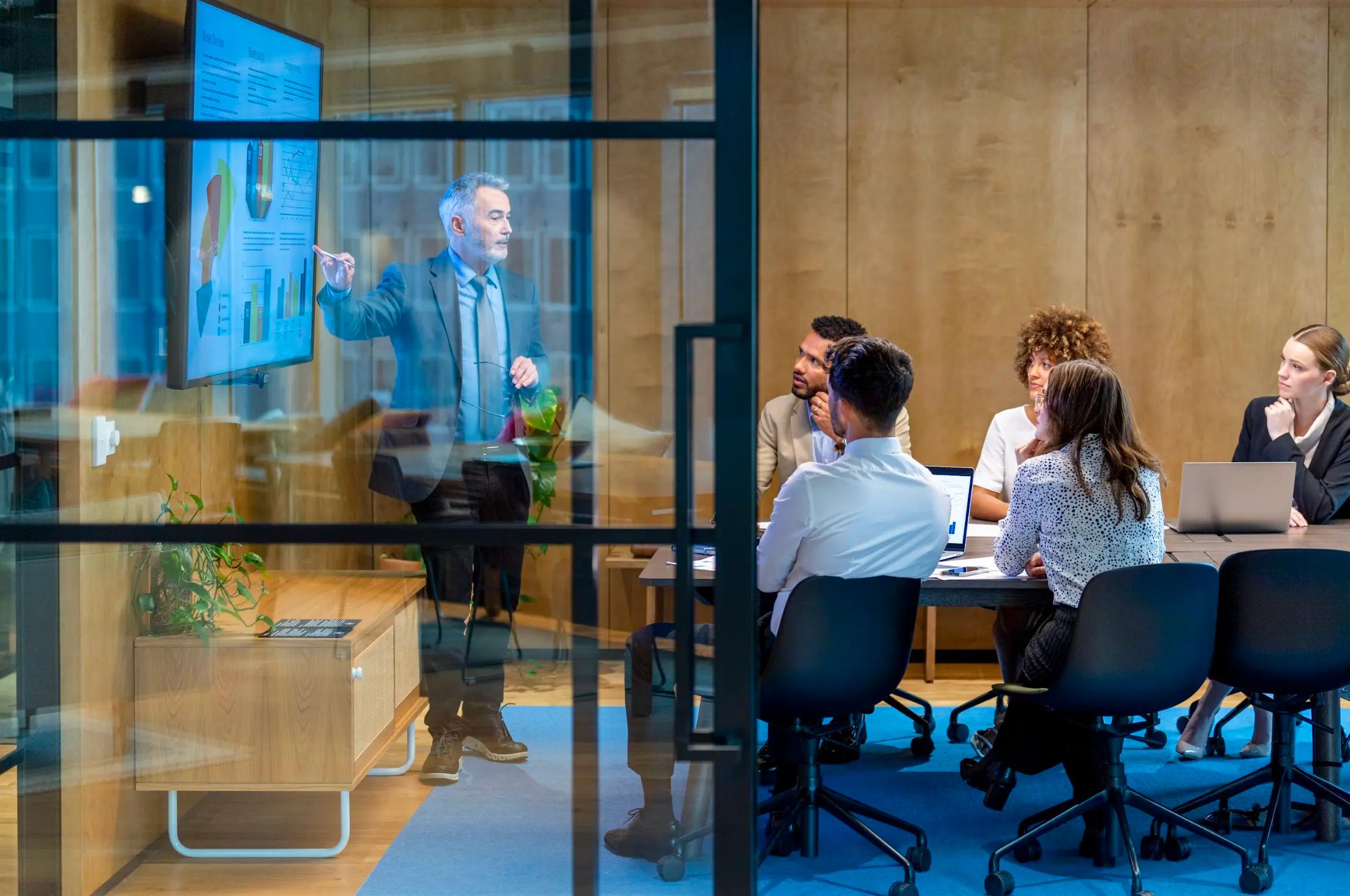 Business professional presenting charts on a large screen to a group seated in a modern glass-walled conference room.